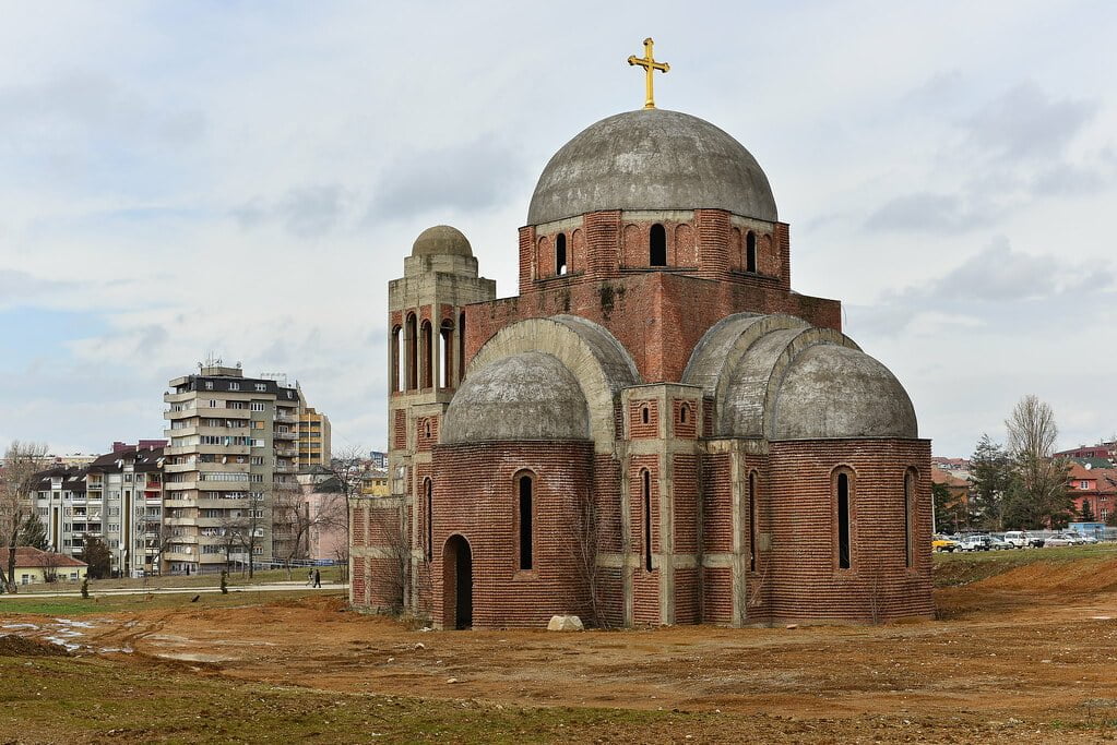 A partially constructed red-brick Orthodox church with a golden cross atop its main dome, surrounded by an undeveloped landscape with residential buildings in the background under a cloudy sky.