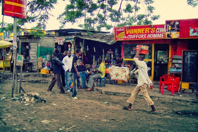 A bustling street scene with people walking by and sitting at a row of colorful makeshift shops and stands under cloudy skies. Signs advertise various services including a barber shop and communication services. A man walks prominently in the foreground carrying a red container on his head.
