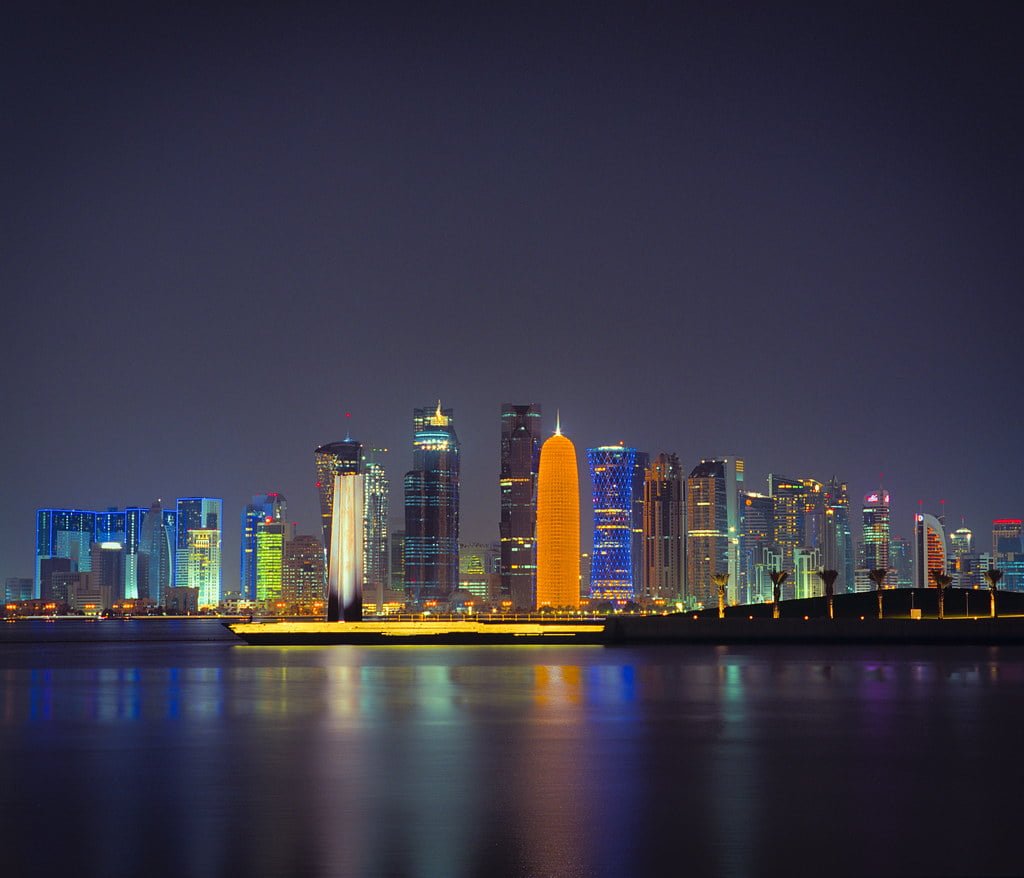 A nighttime cityscape with illuminated skyscrapers reflecting on a calm body of water.