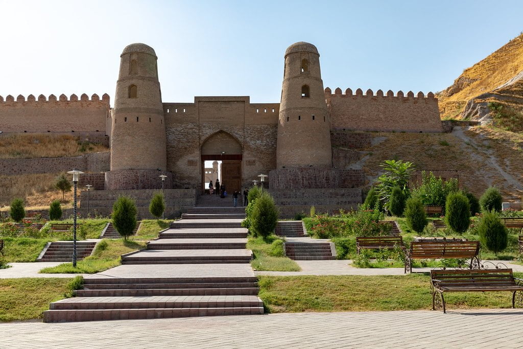 Stone pathway leading to an ancient fortress with twin turrets and arched gateway, surrounded by manicured greenery and benches, under a clear sky.