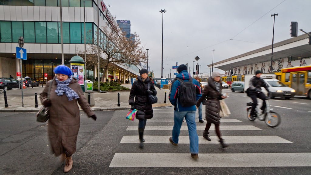 Pedestrians crossing a zebra crossing in a city street with a cyclist passing by and buildings in the background on an overcast day.