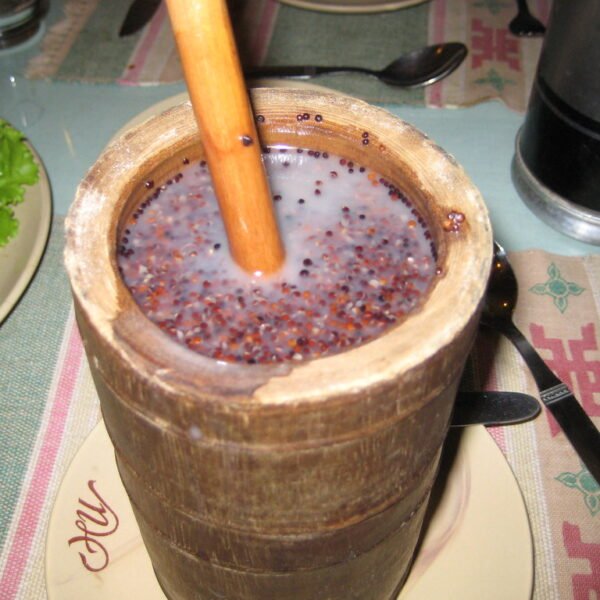A wooden mortar filled with a purple speckled liquid and grains, with a pestle inserted, on a table set with cutlery and partially visible dishes.