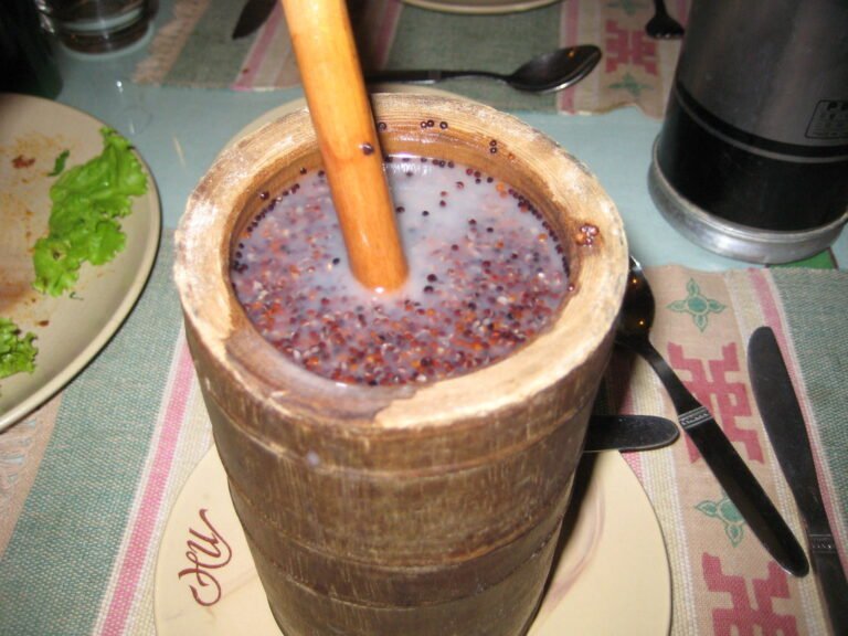 A wooden mortar filled with a purple speckled liquid and grains, with a pestle inserted, on a table set with cutlery and partially visible dishes.