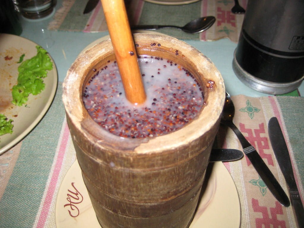 A wooden mortar filled with a purple speckled liquid and grains, with a pestle inserted, on a table set with cutlery and partially visible dishes.