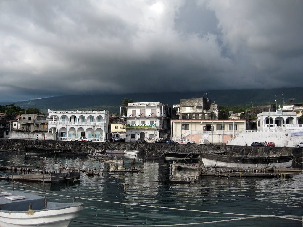 Old waterfront buildings with arched architectural details beside a harbor with docked boats, under a stormy sky with dark clouds.