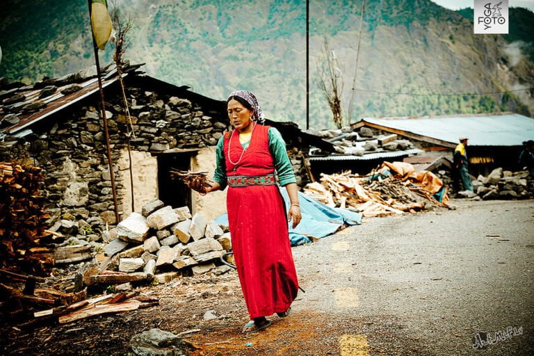 A woman in traditional attire walks through a village carrying firewood, with stone houses and piled wood in the background.
