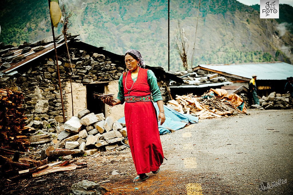 A woman in traditional attire walks through a village carrying firewood, with stone houses and piled wood in the background.