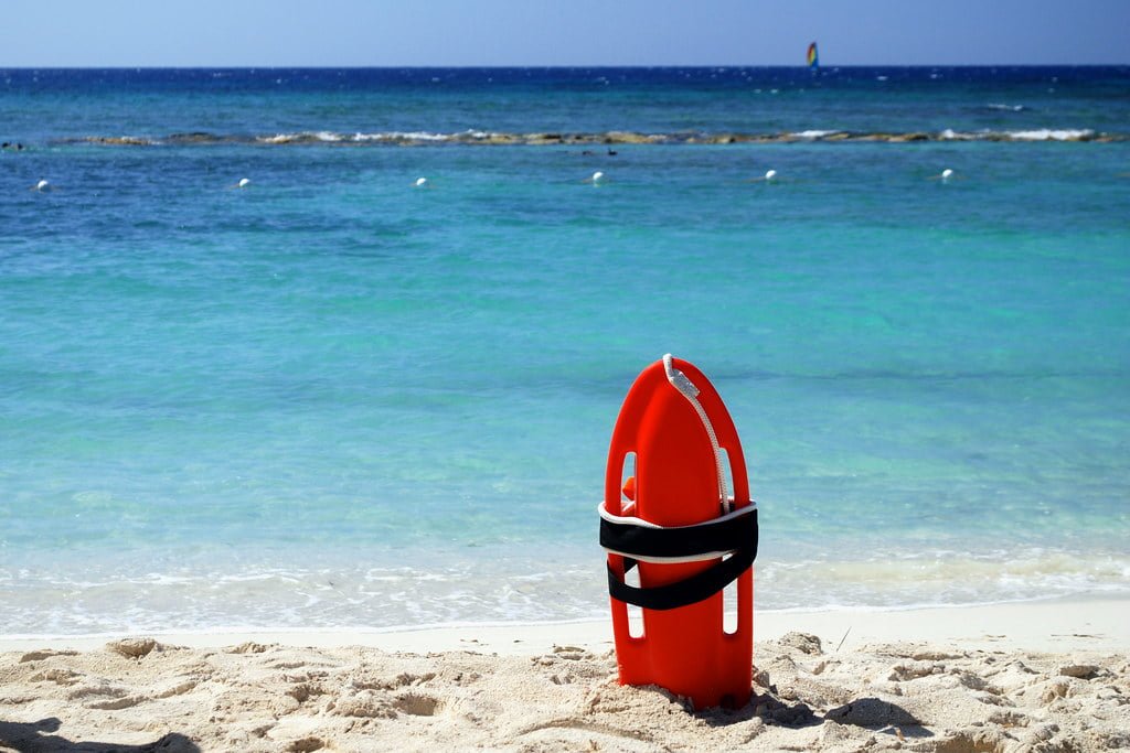 A red rescue buoy stands upright on a sandy beach with clear blue ocean waters in the background and a windsurfer visible in the distance.