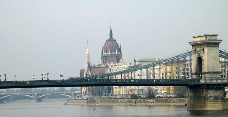 View of the Chain Bridge in Budapest with the Hungarian Parliament Building in the background, over the Danube River on a hazy day.