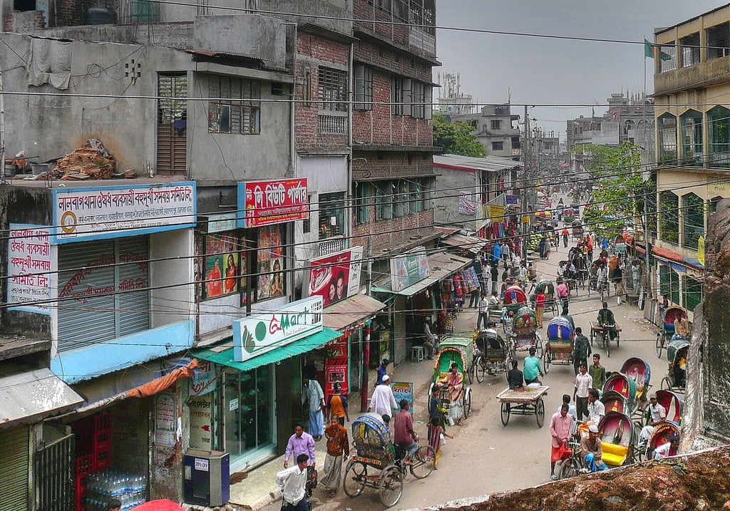 A bustling street scene in a South Asian city with numerous rickshaws and pedestrians, storefronts featuring colorful billboards and signs in a local script, and densely packed multi-story buildings.