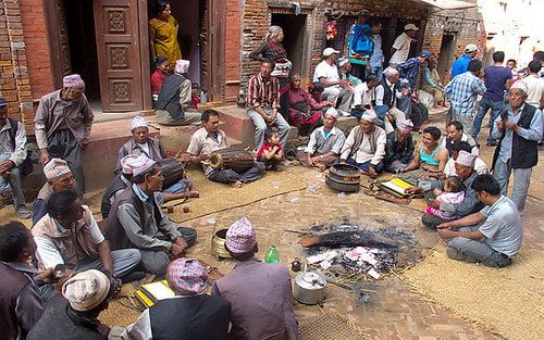 A group of people, many wearing traditional Nepali hats, gathered around a small fire in an outdoor setting with onlookers standing in the background.