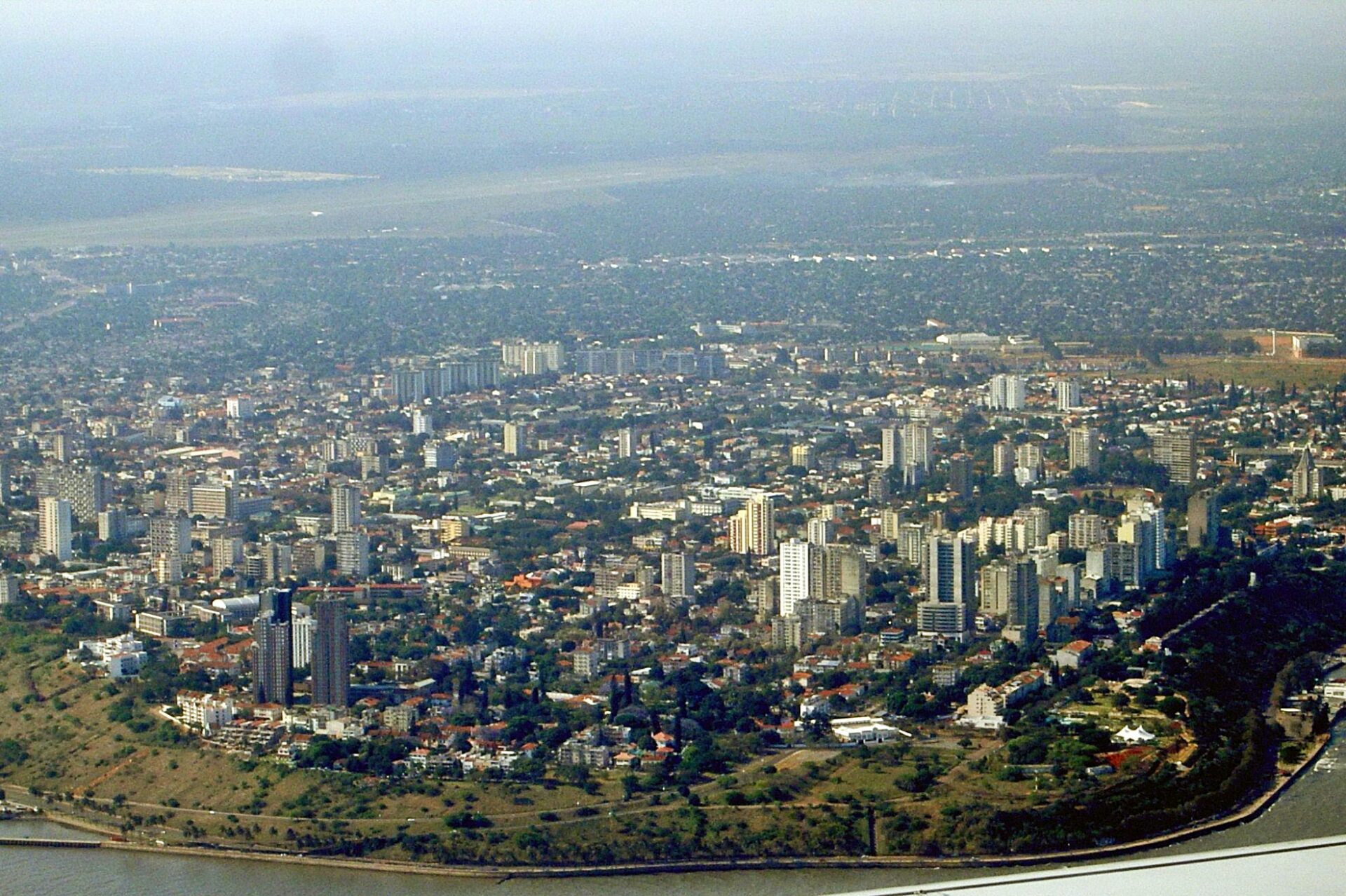 Aerial view of a densely populated cityscape with high-rise buildings near a body of water, under hazy sky conditions.