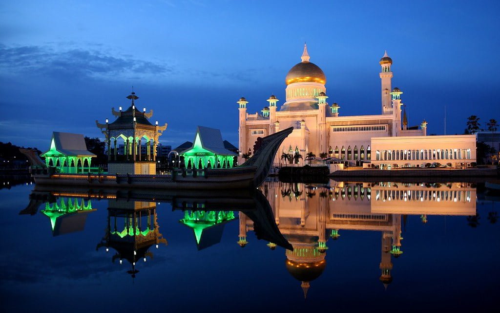 A Sultan Omar Ali Saifuddien Mosque in Brunei, with its golden dome and minarets illuminated, reflected in the calm waters during twilight.