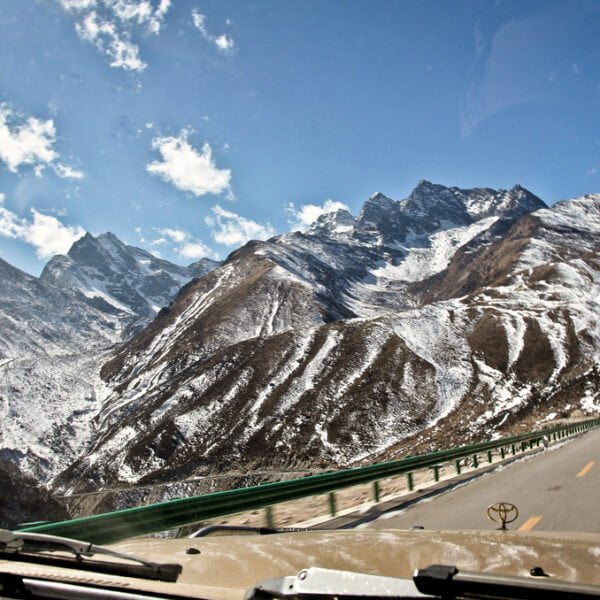 A view through a vehicle's windshield of a winding road leading into snow-capped mountains under a blue sky with scattered clouds.