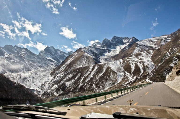 A view through a vehicle's windshield of a winding road leading into snow-capped mountains under a blue sky with scattered clouds.