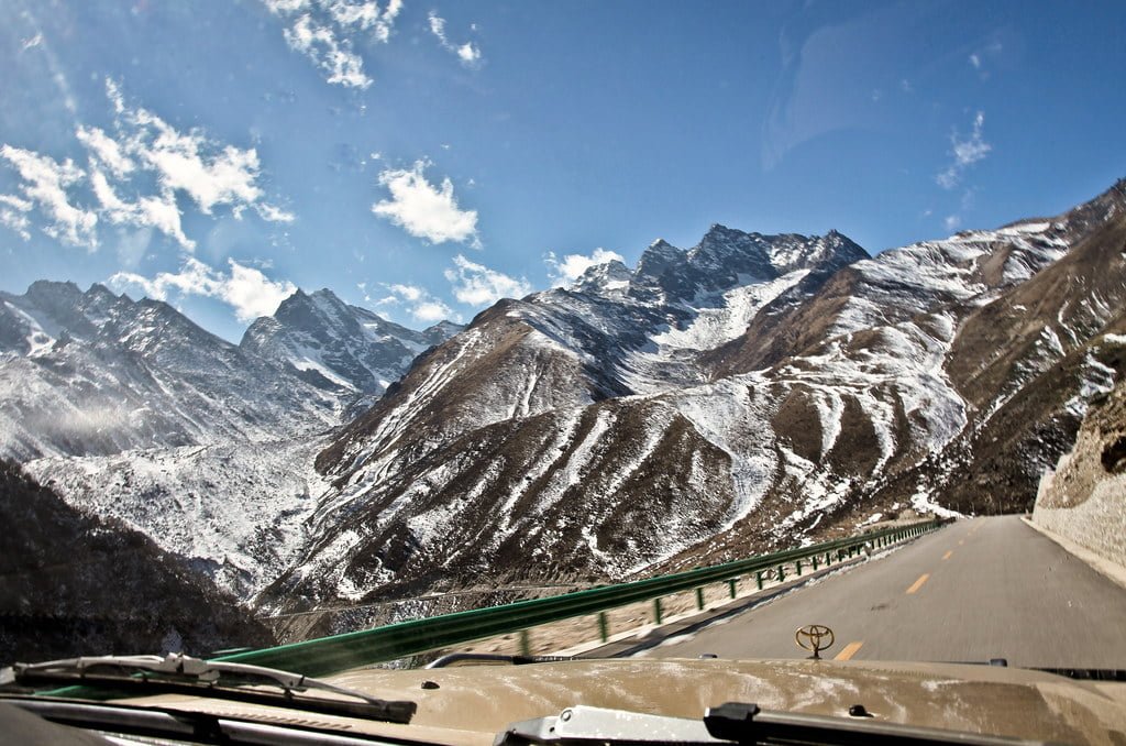 A view through a vehicle's windshield of a winding road leading into snow-capped mountains under a blue sky with scattered clouds.
