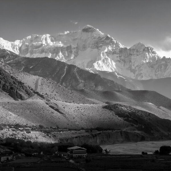 Black and white image of a rugged mountain landscape with a small building in the foreground and towering snow-covered peaks in the background, under a clear sky.