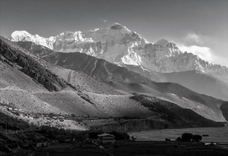 Black and white image of a rugged mountain landscape with a small building in the foreground and towering snow-covered peaks in the background, under a clear sky.