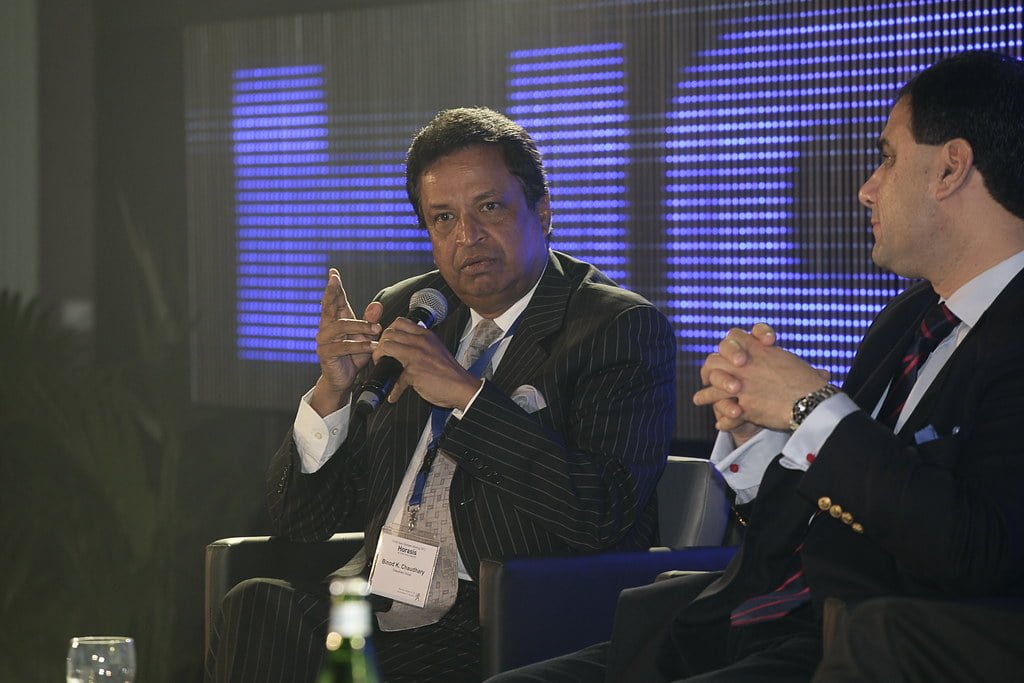 Two men in business suits sitting at a conference event, one with a microphone in hand, with a blue LED screen in the background.