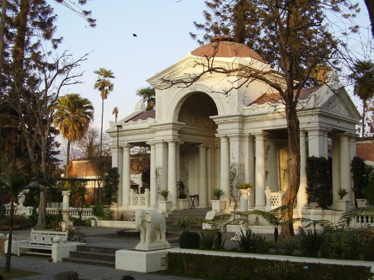 Alt text: An elegant white pavilion with classical columns and arches surrounded by a lush garden with palm trees, statues, and benches in a serene park setting. A bird flies in the clear sky above.