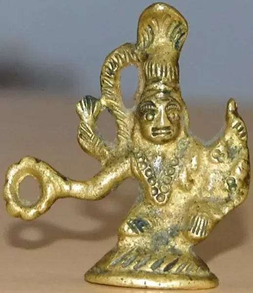 A small, gold-colored figurine with Hindu deity features holding a ring or object in one hand, and the other hand raised, set against a blurry background.