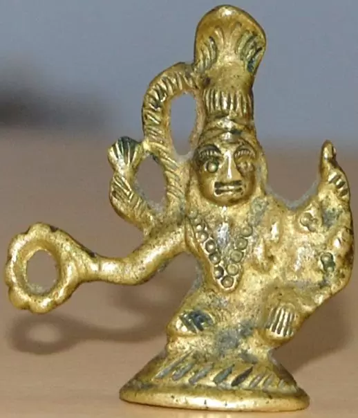 A small, gold-colored figurine with Hindu deity features holding a ring or object in one hand, and the other hand raised, set against a blurry background.