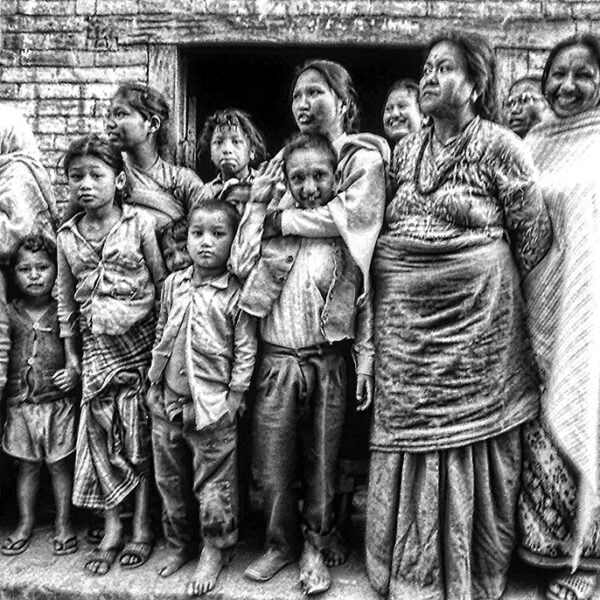 A group of people from various generations, predominantly women and children, stand closely together in front of a brick wall, exhibiting a range of emotions. The image is in grainy black and white, adding to the intensity of the scene.