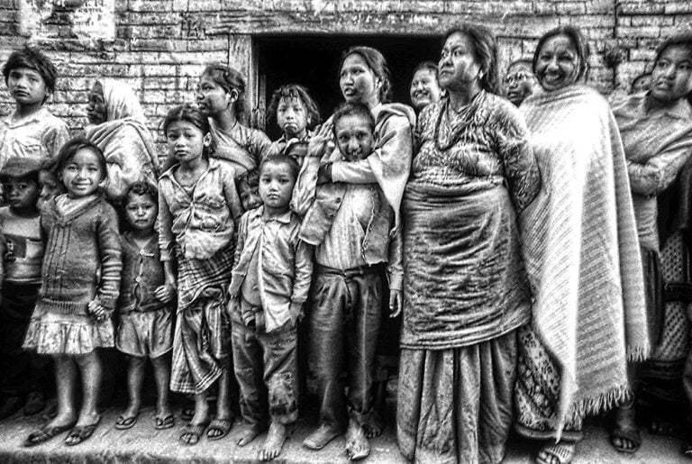 A group of people from various generations, predominantly women and children, stand closely together in front of a brick wall, exhibiting a range of emotions. The image is in grainy black and white, adding to the intensity of the scene.