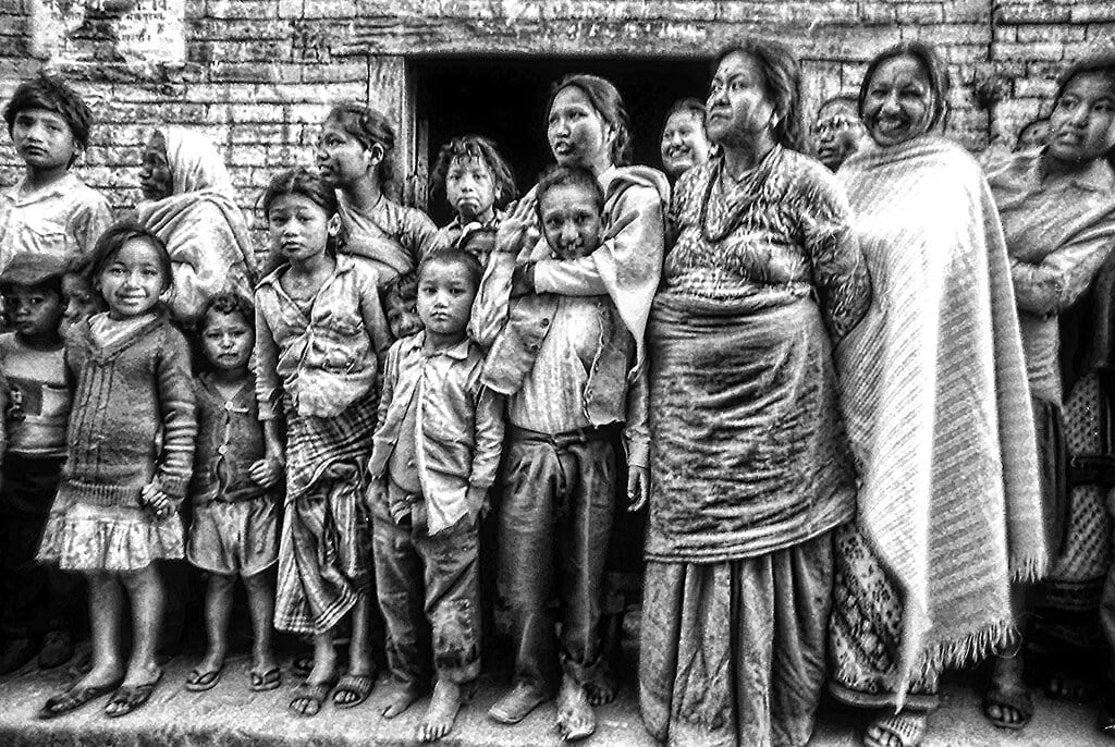 A group of people from various generations, predominantly women and children, stand closely together in front of a brick wall, exhibiting a range of emotions. The image is in grainy black and white, adding to the intensity of the scene.