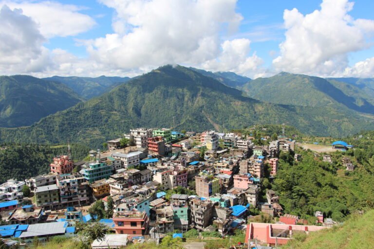 A mountain town with multicolored buildings nestled on terraced slopes, surrounded by lush green hills under a blue sky with scattered clouds.