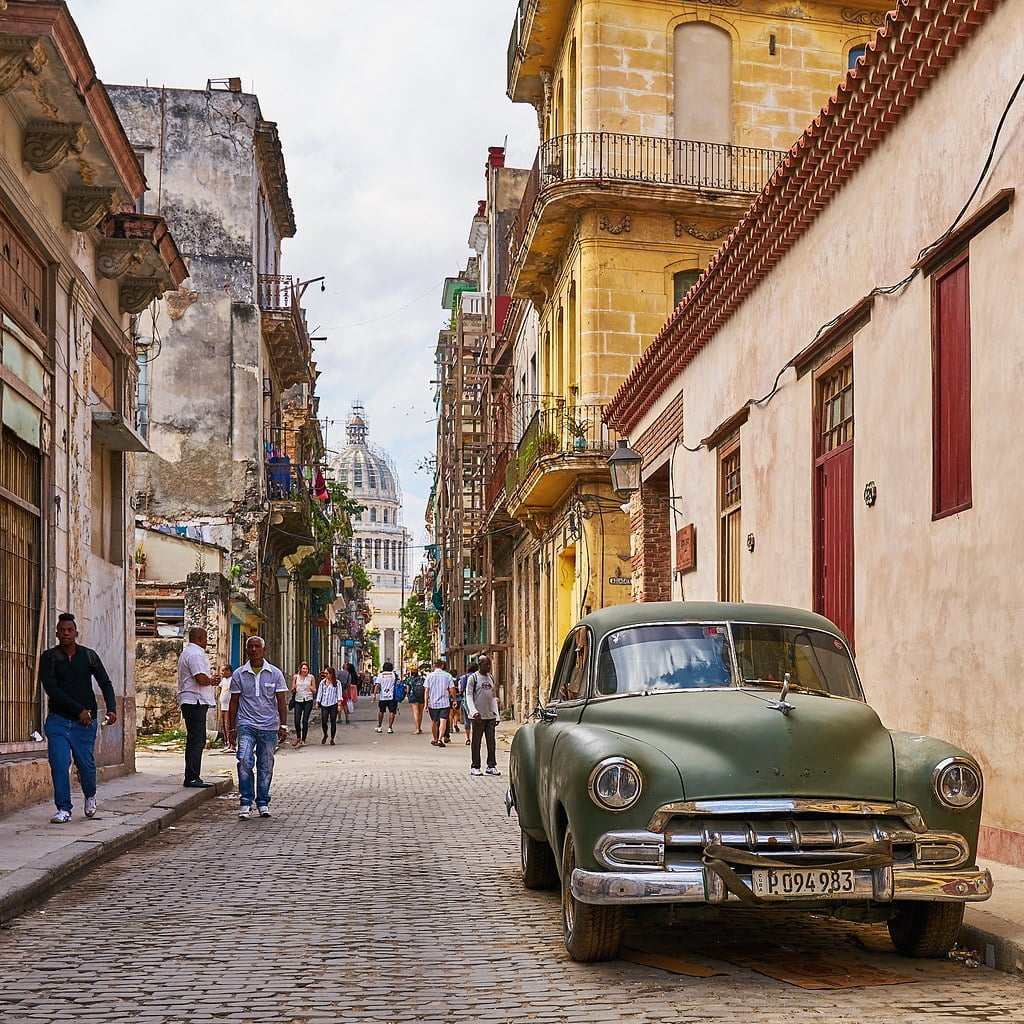 Alt text: A vintage green car parked on a cobblestone street with pedestrians walking by, surrounded by old buildings, with a large domed building in the background.