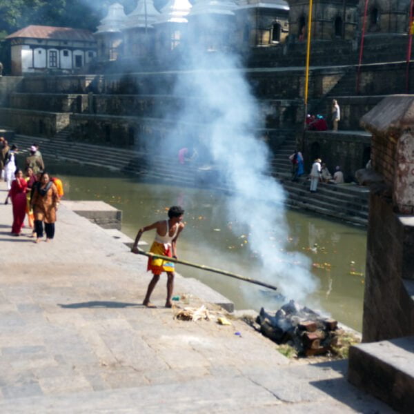 A person is tending to a fire by a stepped water tank near a temple complex, with smoke rising, as several other individuals, some in traditional attire, walk along the temple stairs.