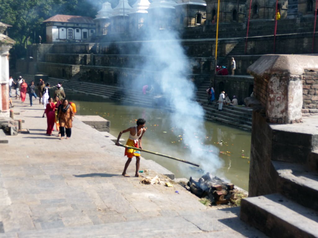 A person is tending to a fire by a stepped water tank near a temple complex, with smoke rising, as several other individuals, some in traditional attire, walk along the temple stairs.