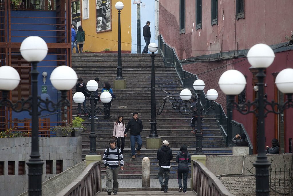 A bustling urban staircase with individuals walking and conversing, flanked by red and yellow buildings, and ornate lampposts with spherical white lights in the foreground.