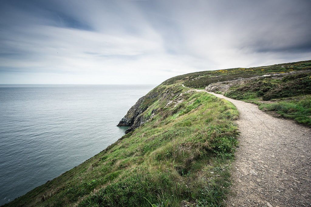 Coastal path along a cliff with overcast skies, overlooking a serene ocean.