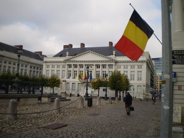 A Belgian flag hangs prominently in front of a classic European building with white façade and grey roof under an overcast sky. A cobblestone square with pedestrians and trees in planters leads to the building.