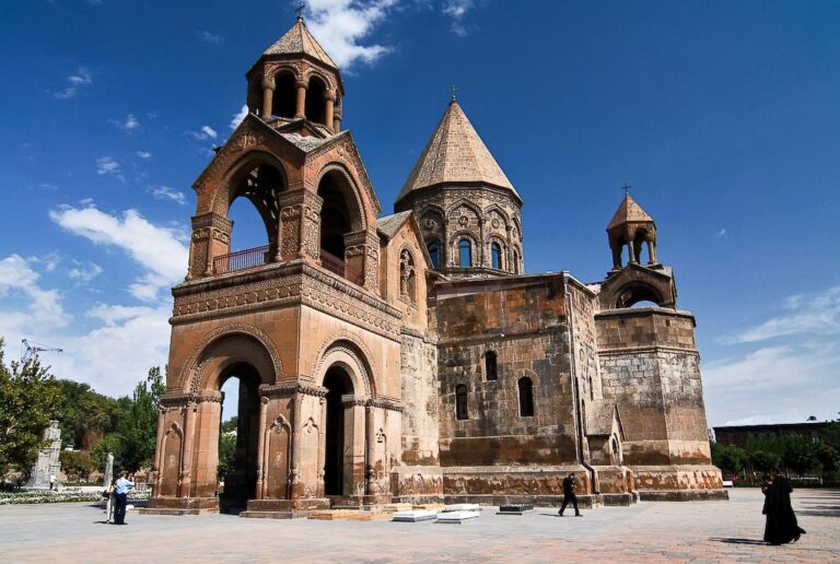 An ancient stone church with a bell tower and domes under a blue sky, with people walking around the sunny courtyard.