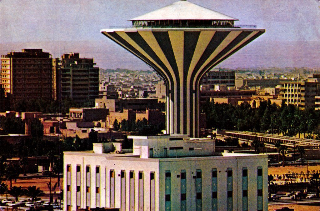 A vintage postcard image featuring a cityscape with a distinctive flying saucer-shaped building on a large pedestal dominating the foreground, surrounded by mid-rise buildings and a clear sky.