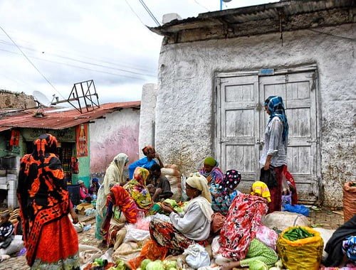 A group of people in colorful traditional attire sitting in a circle on the ground, possibly engaged in an activity or work, in a rural setting with an old white building with a closed door in the background.