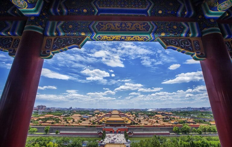 A panoramic view of the Forbidden City in Beijing framed by ornate traditional Chinese architectural details under a blue sky with scattered clouds.