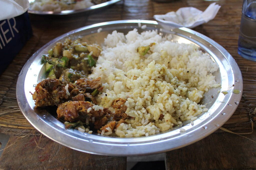 A stainless steel plate filled with white rice, spicy chicken pieces, and cooked vegetables, placed on a rustic wooden surface, with a water glass and napkin in the background.