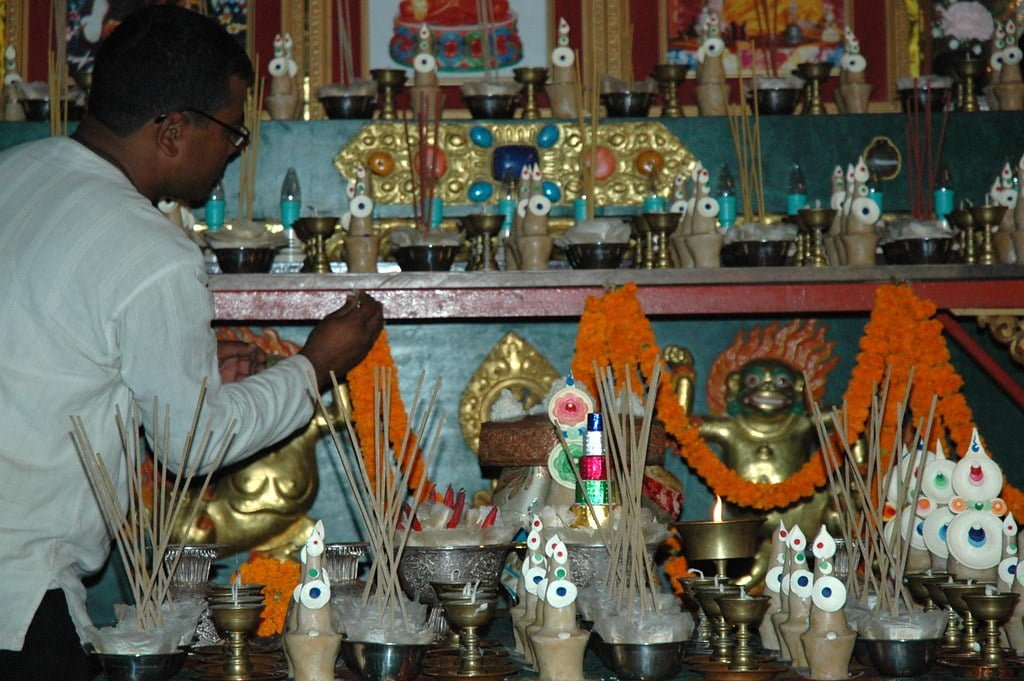 A person lighting incense sticks at an altar adorned with marigold flowers, Buddhist statuettes, and various ritual items.