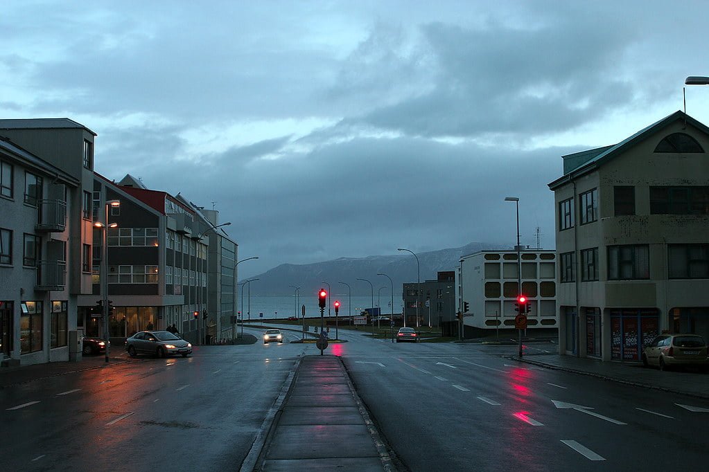 A town street at dusk with wet roads reflecting the red traffic signal, cars driving, and mountains visible in the background under a cloudy sky.