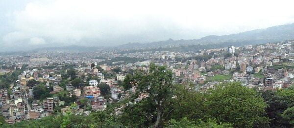 Panoramic view of a densely populated city with buildings spread across a hilly terrain, partially obscured by haze, with green foliage in the foreground.