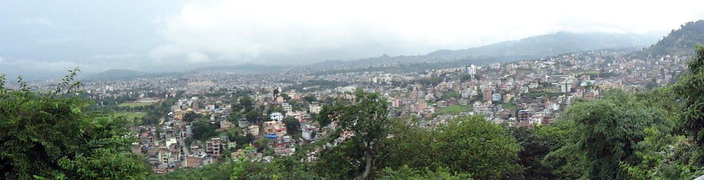 Panoramic view of a densely populated city with buildings spread across a hilly terrain, partially obscured by haze, with green foliage in the foreground.