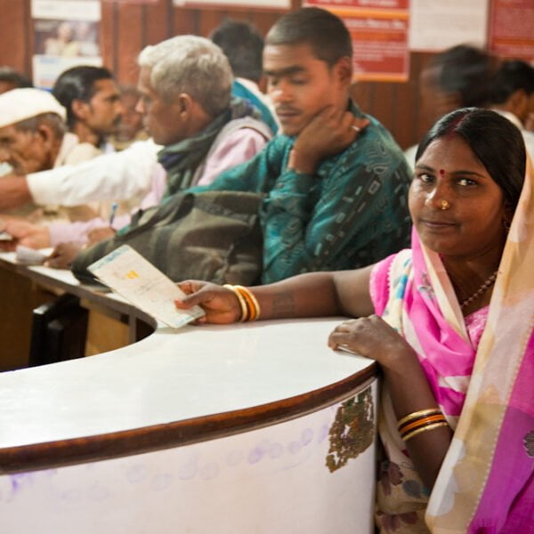 A woman in a colorful saree stands at a counter with a document in a busy Indian post office, with other customers in the background engaging with postal workers.