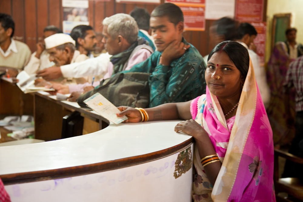 A woman in a colorful saree stands at a counter with a document in a busy Indian post office, with other customers in the background engaging with postal workers.