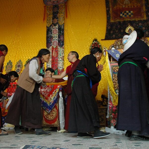 Tibetan people in traditional attire participating in a cultural ceremony with monks wearing red robes, inside a room with vibrant yellow walls and decorative textiles.