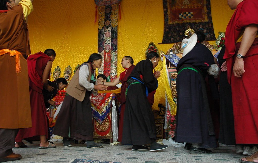 Tibetan people in traditional attire participating in a cultural ceremony with monks wearing red robes, inside a room with vibrant yellow walls and decorative textiles.