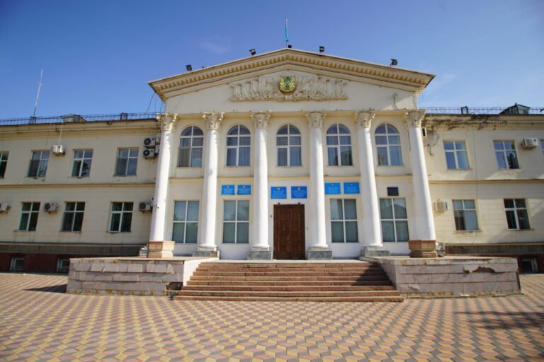 A photo of a neoclassical government building with white columns, a central entrance with wooden doors, and steps leading up to it, under a clear blue sky.